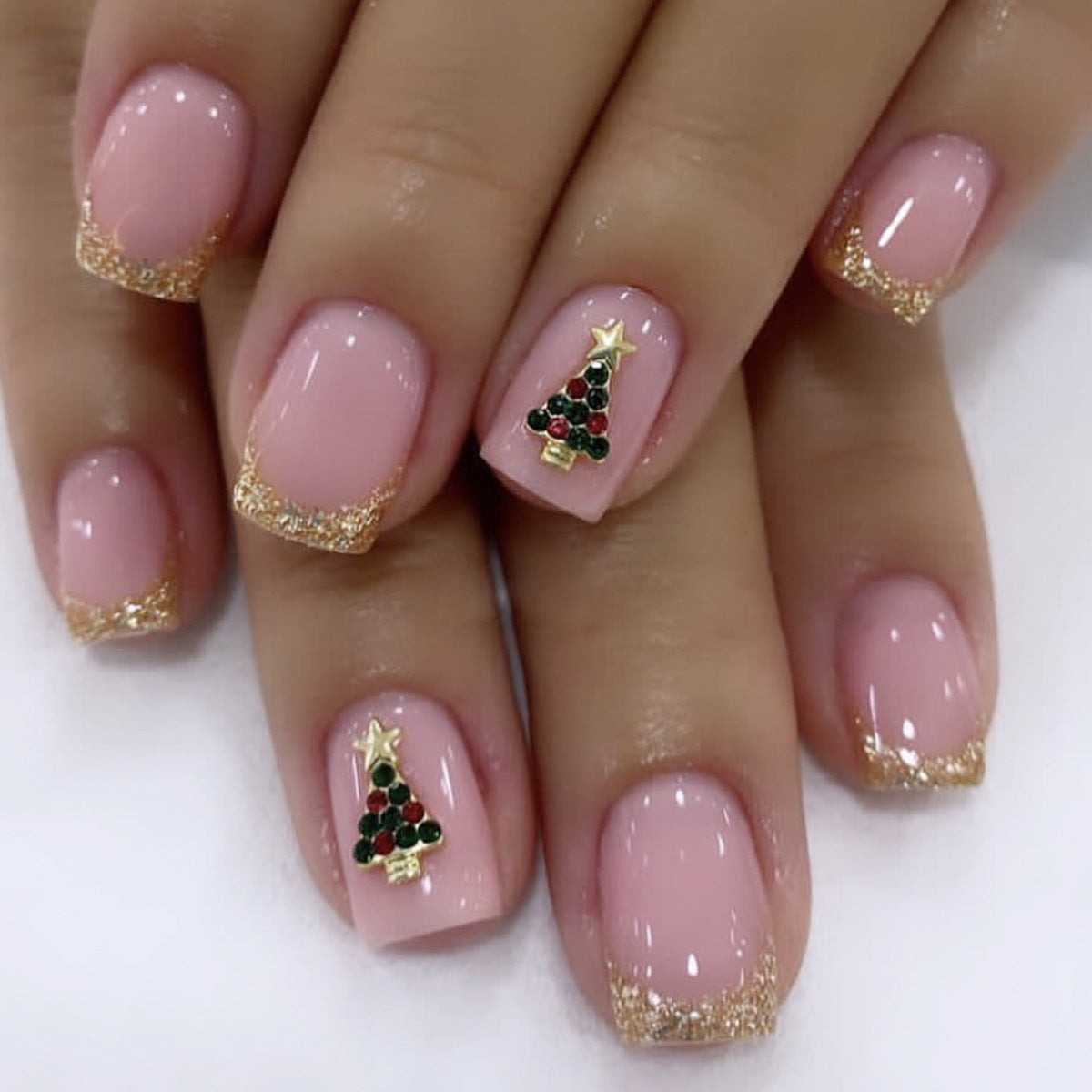 A set of press-on nails featuring a pink base with gold glitter and a Christmas tree design, displayed on a hand.