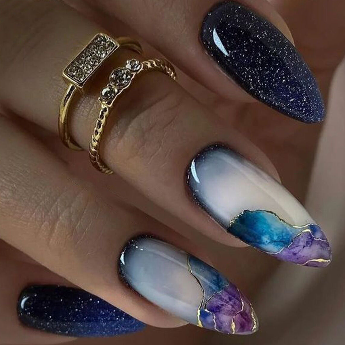 Nail art with blue, white, and purple design on a hand wearing gold rings.