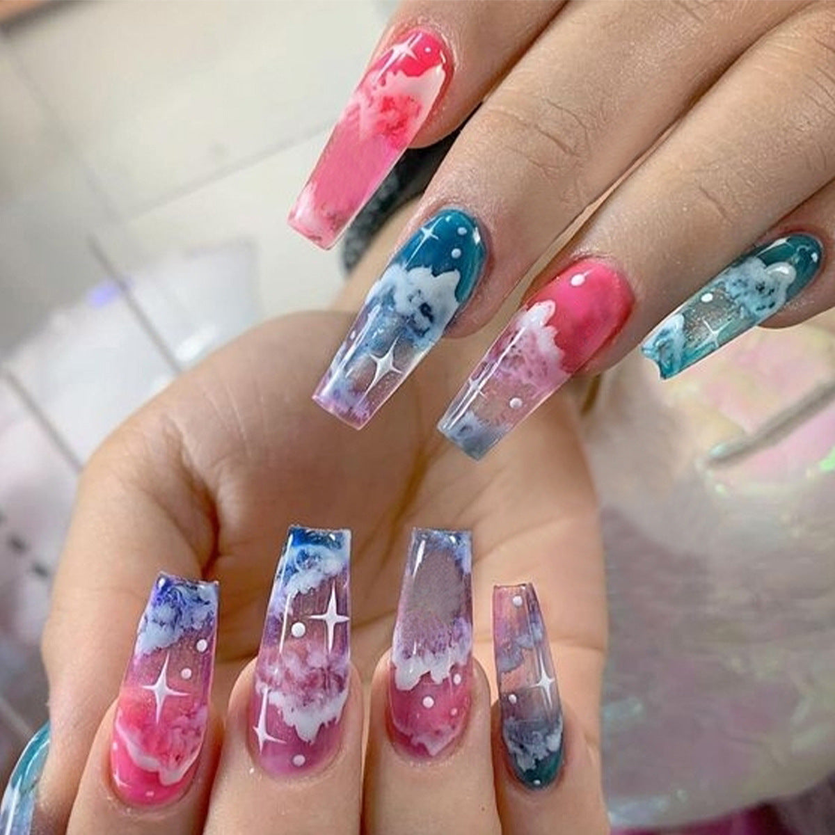 A set of press-on nails featuring an ombre design with pink, blue, and white colors, and glitter accents.