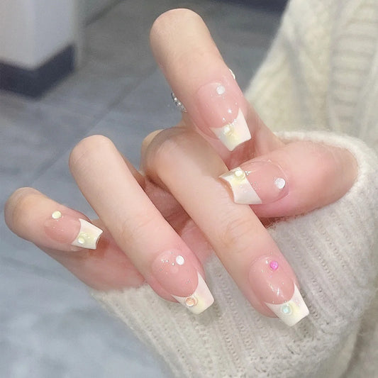 A set of beige press-on nails with rhinestone accents, presented on a hand with a sweater in the background.