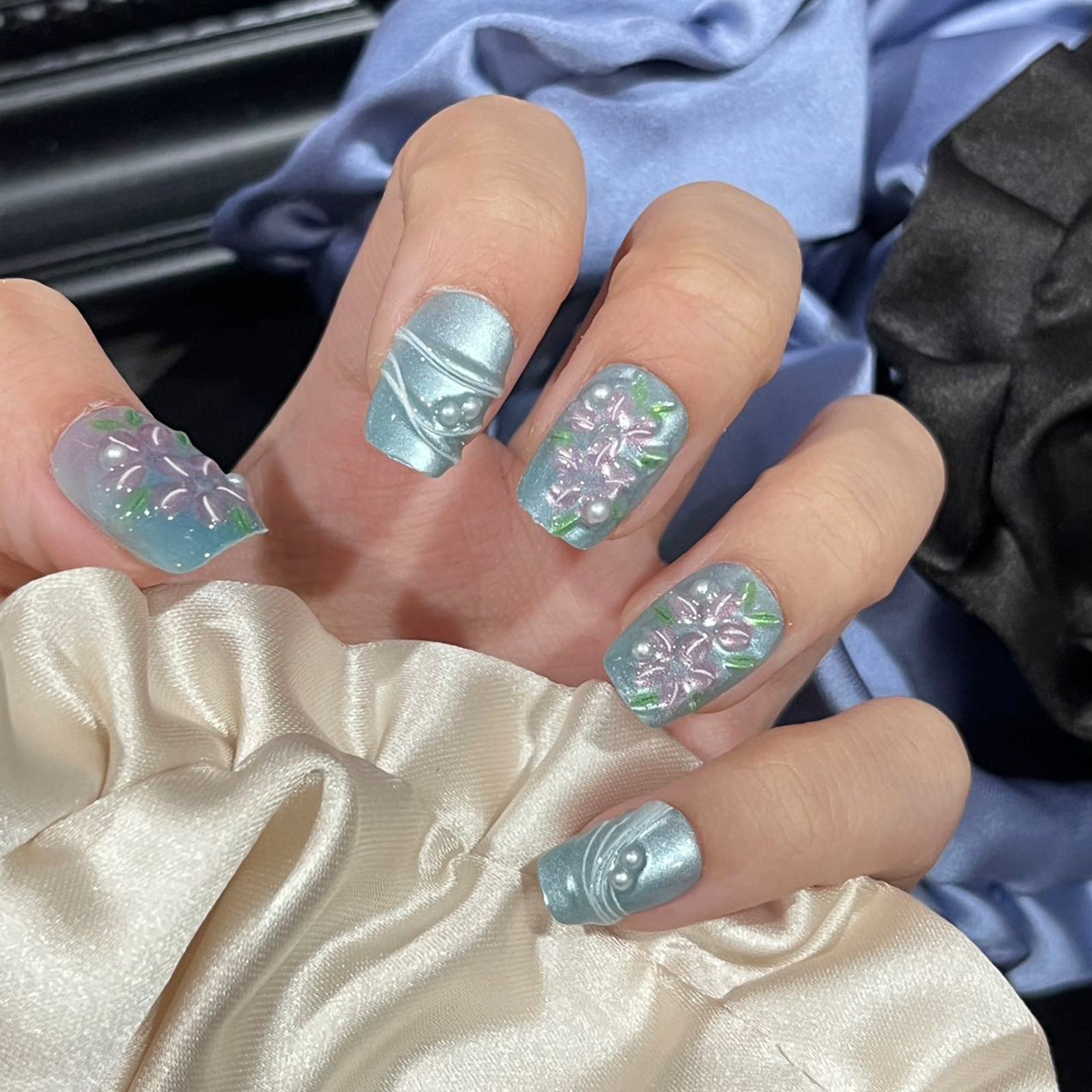 A set of short square press-on nails with blue floral designs and shimmering pearl accents, displayed on a hand against a soft fabric background.