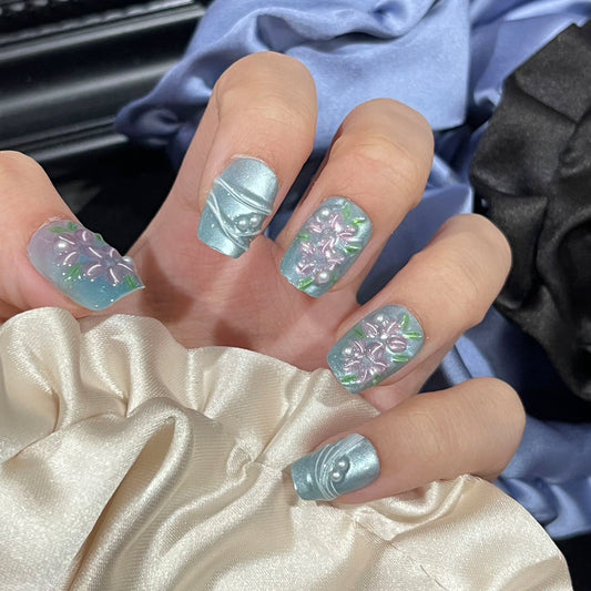 A set of short square press-on nails with blue floral designs and shimmering pearl accents, displayed on a hand against a soft fabric background.