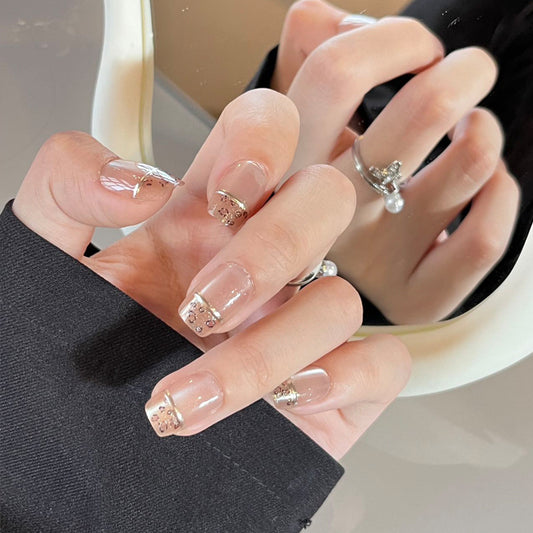 A set of short square clear nails with sparkling gold accents, displayed on a person's hands.
