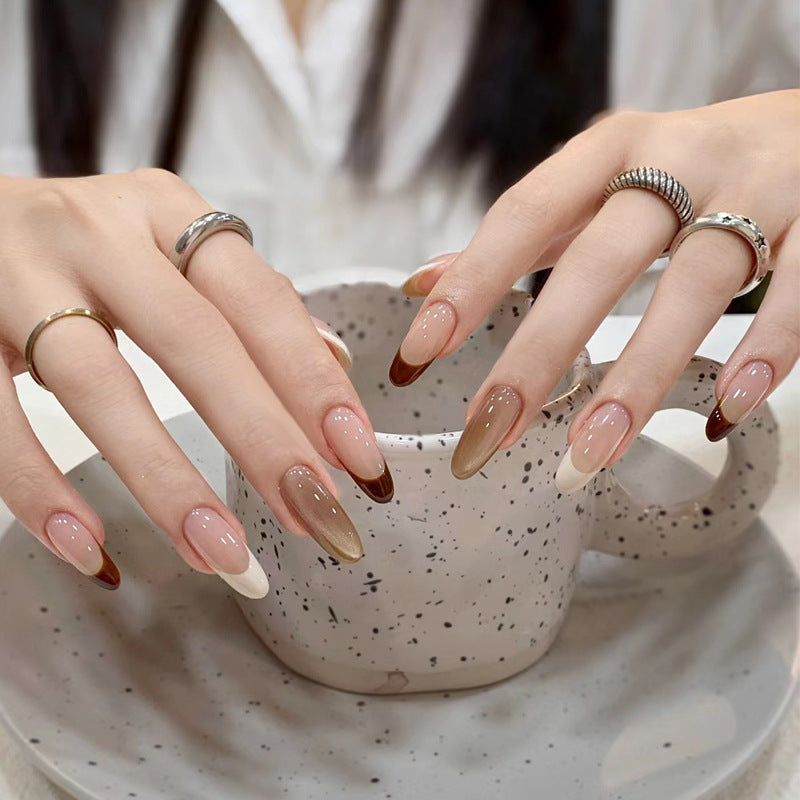A set of long, almond-shaped, nuanced brown press-on nails with a glossy finish, being worn by a person with a cup of coffee in the background.