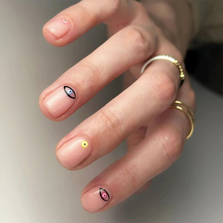A set of beige press-on nails with short oval shapes and eye design accents, displayed on a person's hand.