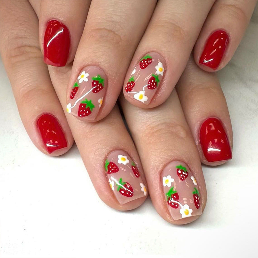 A set of short square red press-on nails with strawberry and flower accents, shown on a person's nails.