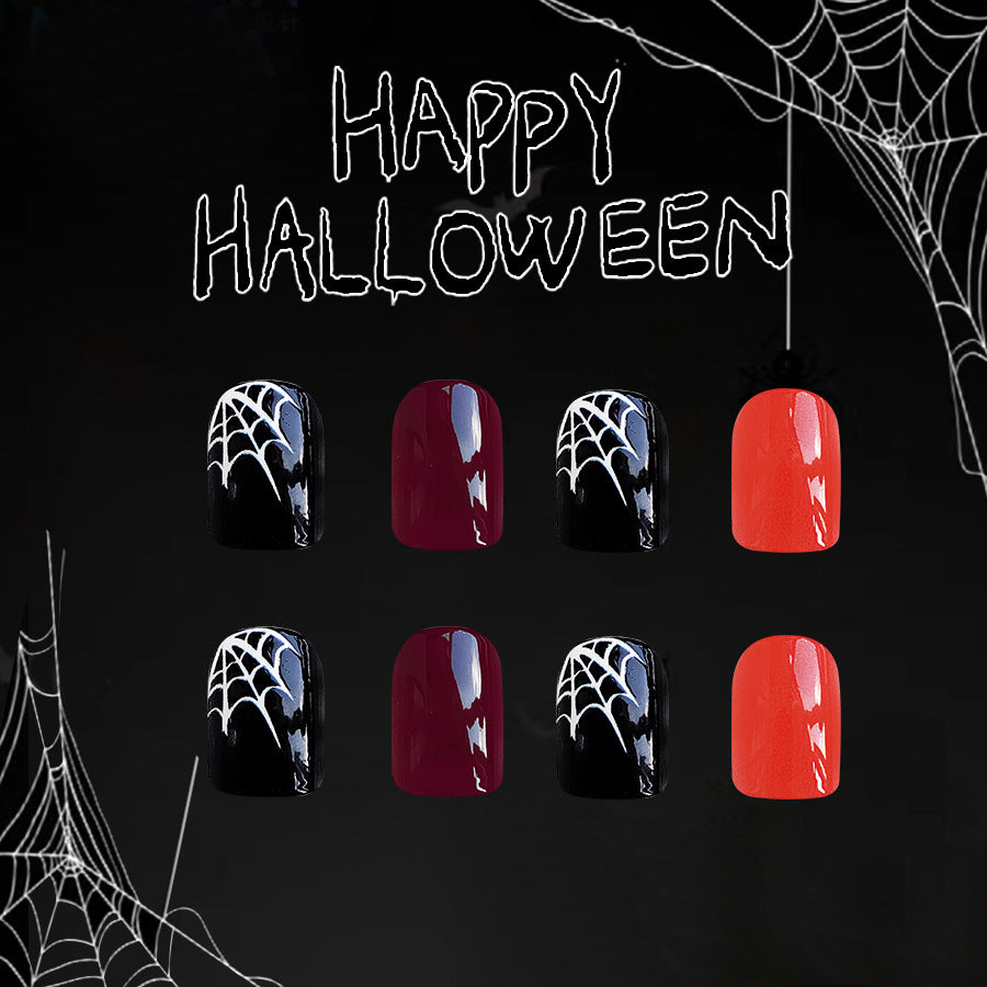 A set of press-on nails displayed in a Halloween-themed setting with spider webs and the words 'HAPPY HALLOWEEN' written above. The nails have a short square shape and come in black and red colors with a cobweb design.