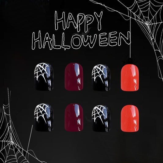A set of press-on nails displayed in a Halloween-themed setting with spider webs and the words 'HAPPY HALLOWEEN' written above. The nails have a short square shape and come in black and red colors with a cobweb design.