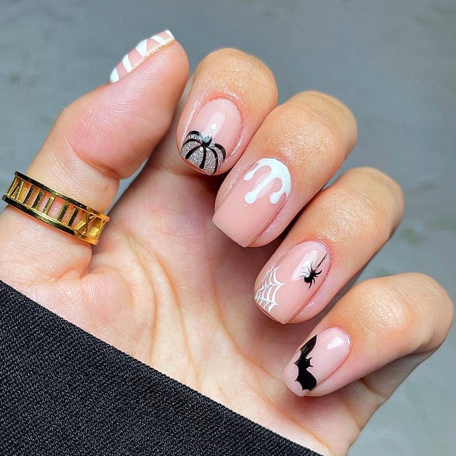 A set of short square pink press-on nails with various Halloween accents, including bats, ghosts, and the letter 'M' in black.