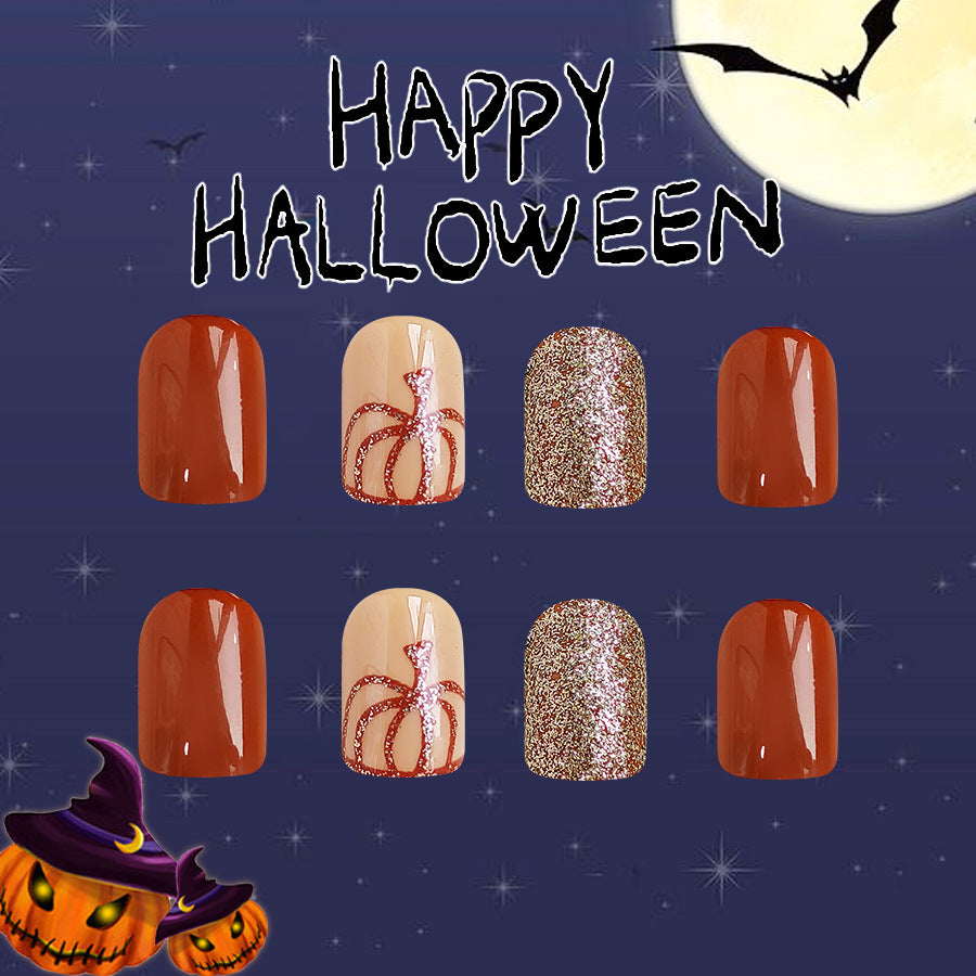 A set of four press-on nails with a Halloween theme. The nails are painted in shades of orange and beige with glitter accents, and feature decorative designs including a spider and a pumpkin.