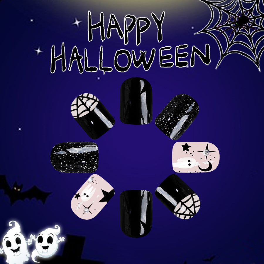 A set of short square black press-on nails with glitter accents and a playful ghost design, displayed against a Halloween-themed background with a spider web, pumpkins, and bats.