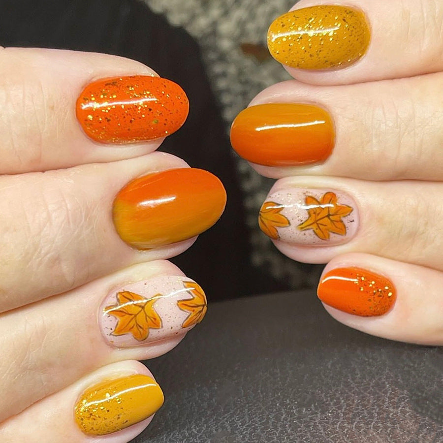 A set of press-on nails featuring a combination of orange, yellow, and glitter, with leaf accents designed to resemble autumn leaves.