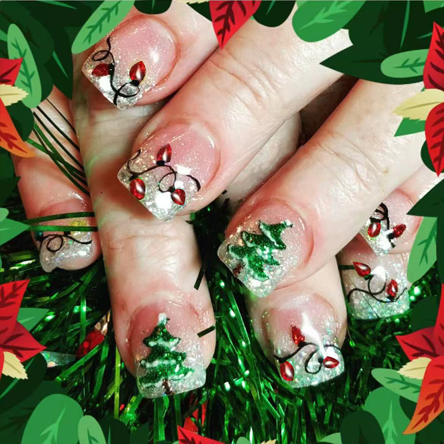 A set of press-on nails featuring a glittering silver design with holiday trees and lights, displayed on a hand against a green and red leafy background.