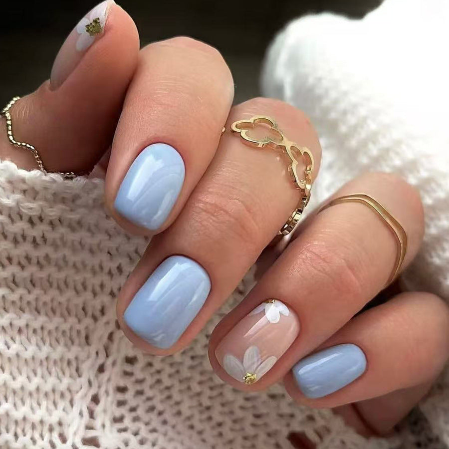 Hand with light blue nails and floral designs, wearing gold rings, against a textured white background.