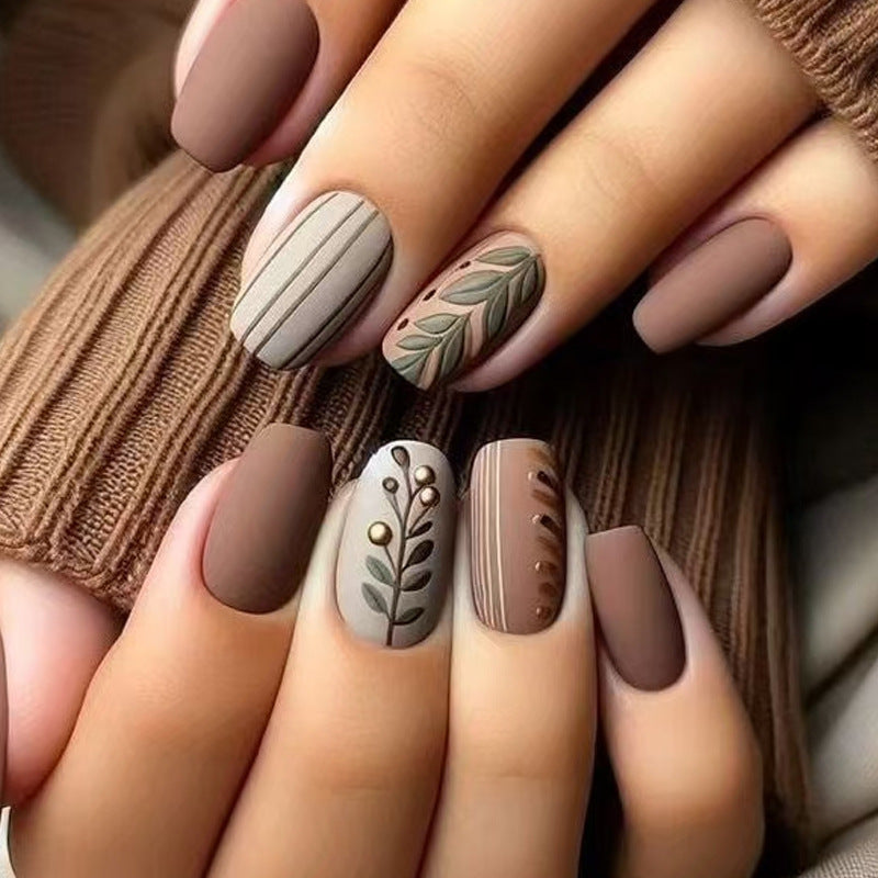 A set of medium square matte nails with brown color and botanical accents, such as leaves and stripes.