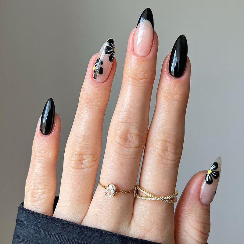 A set of press-on nails featuring black and clear floral accents on an almond nail shape displayed on a hand.