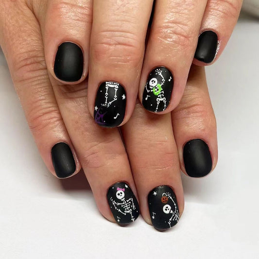 A set of black press-on nails with a skeleton and Halloween design, displayed on a hand.