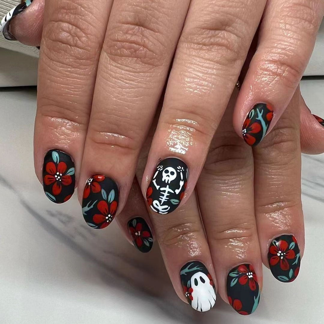 A set of press-on nails featuring a black, red, and white color scheme with decorations of skeletons, flowers, and the word 'BOO!' displayed on a person's hands.