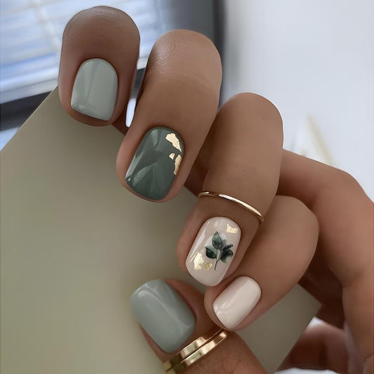 A set of press-on nails featuring sage green and gold leaf accents, with a floral design on the gold leaf-accented nail.