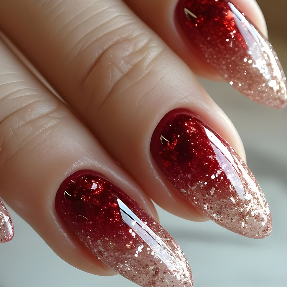 A set of burgundy press-on nails with glittering ombre finish, displayed on a hand.