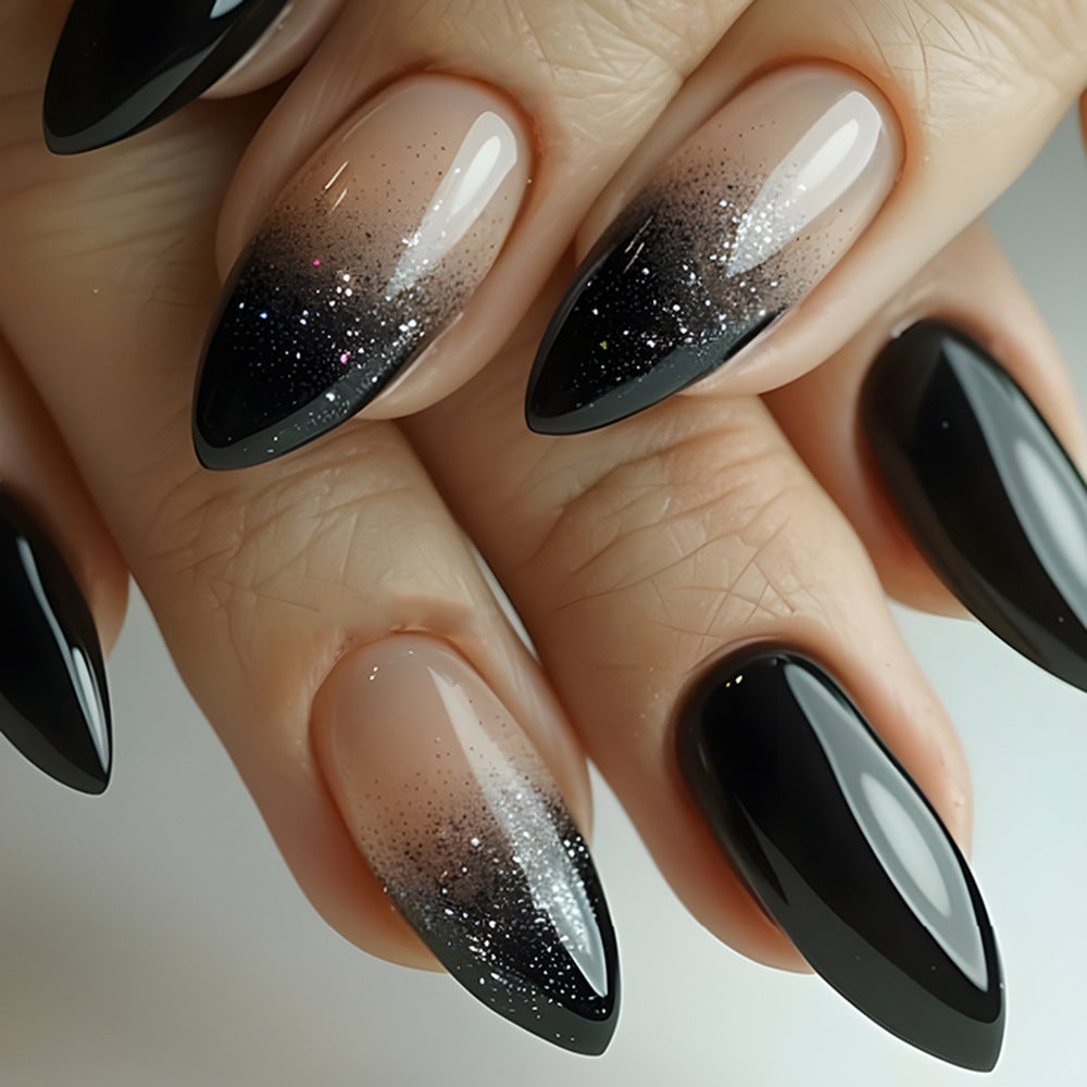 A close-up image of a person's fingernails, featuring press-on nails with a gradient design from beige to black with glitter throughout.