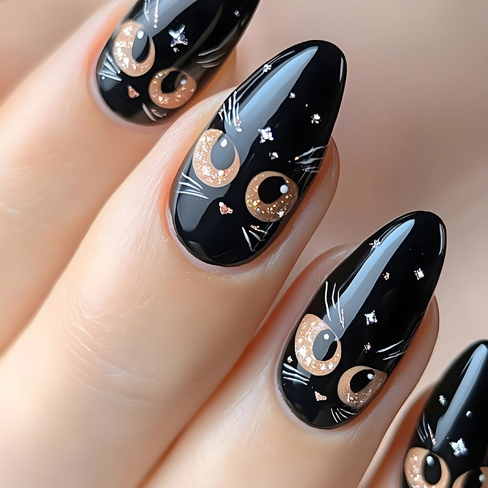 A set of almond-shaped black nails with a glossy finish, decorated with white and gold cat face patterns and sparkling glitter details.