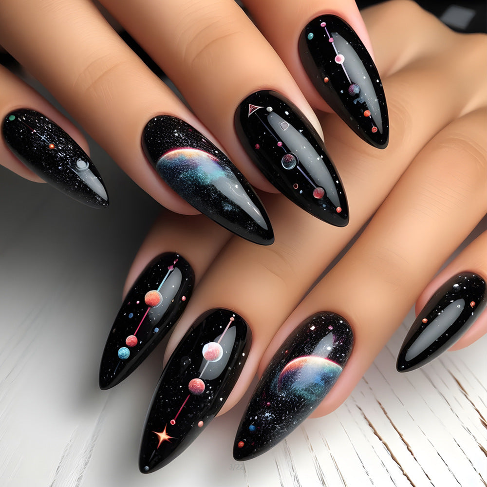 A set of long almond-shaped black nails with cosmic planetary designs, including stars and planets.