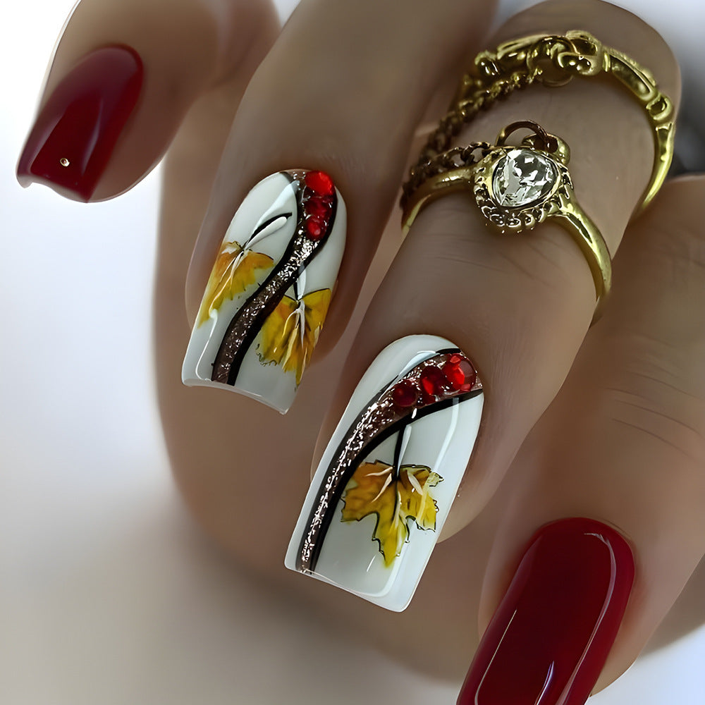 A set of long square white and yellow press on nail sets with glittering leaf design and red accents, displayed on a hand with red nail polish and a golden ring.