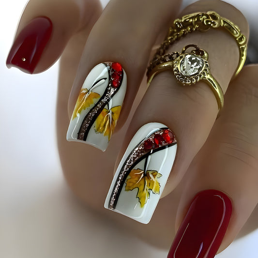 A set of long square white and yellow press on nail sets with glittering leaf design and red accents, displayed on a hand with red nail polish and a golden ring.