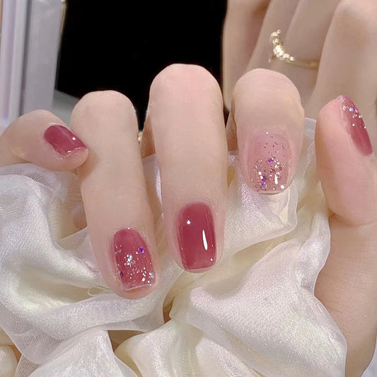 A set of press-on nails featuring a burgundy color with glitter accents, displayed on a person's hand against a white fluffy background.