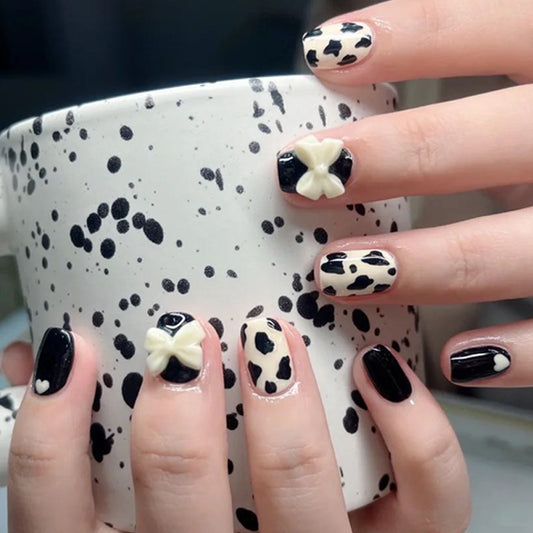 A set of short-length square press-on nails featuring a black and white animal print with 3D bow accents, displayed on a person's hands holding a white cup.