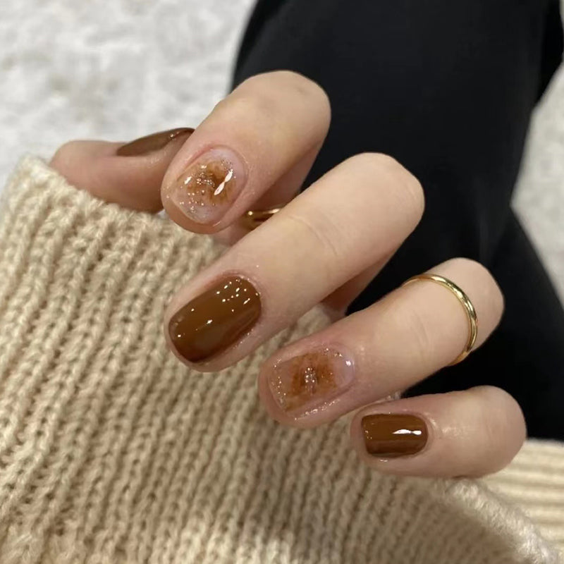 A pair of chocolate brown press-on nails with sparkling accents, shown resting on a person's hands.