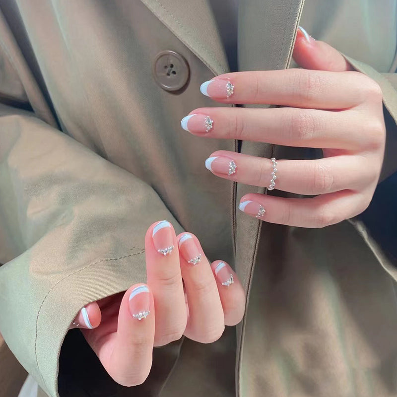 A person's hand with baby pink press-on nails and rhinestone accents, partially covered by a beige jacket.