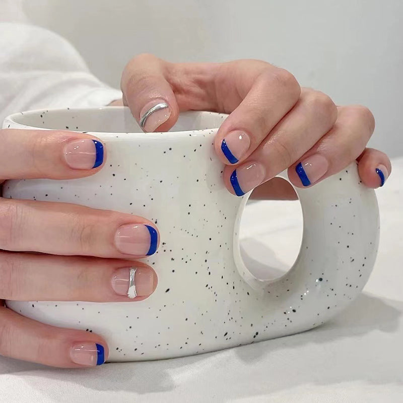 A set of press-on nails featuring a French tip design in beige and sapphire blue with a silver stripe accent, shown resting on a white cup.