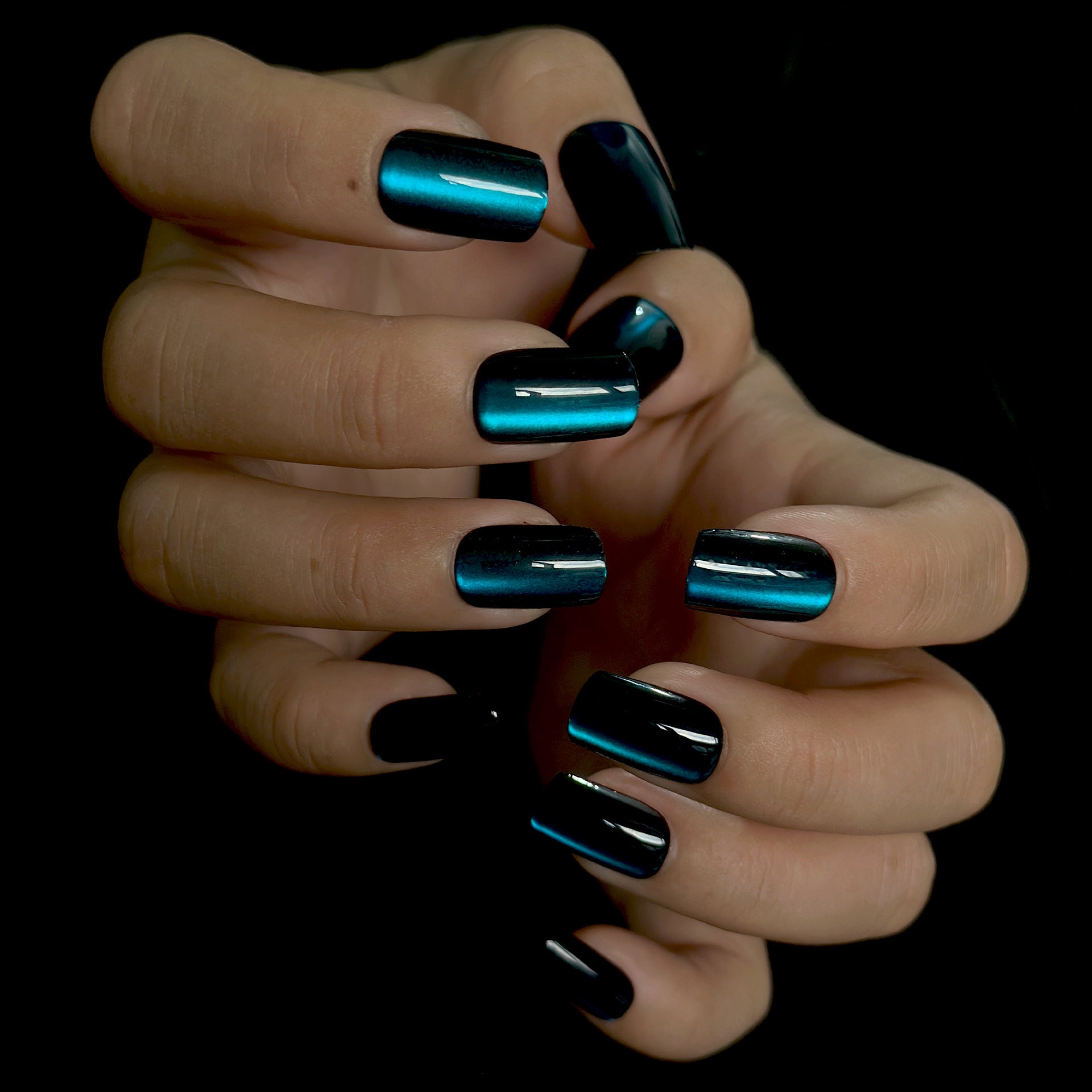 A hand showcasing press on nails with a long square shape. The nails are painted in black and teal colors with an iridescent finish.