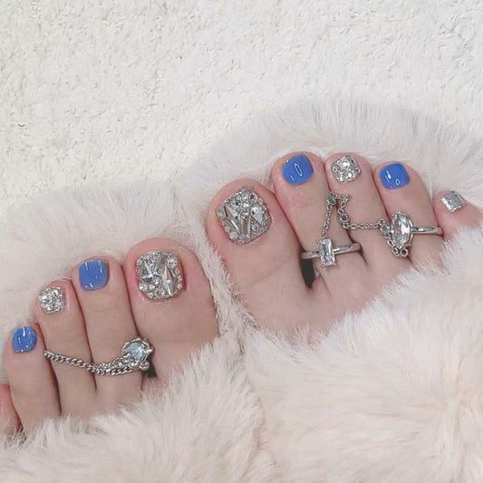 A set of blue false nails with rhinestone embellishments, displayed on a foot resting on a fluffy white surface.
