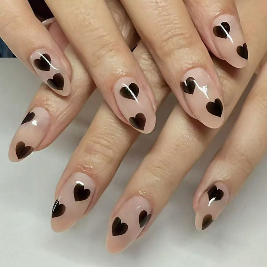 A photo showing a set of manicured nails with a short almond shape, painted in a transparent pink color, each adorned with a small black heart design.