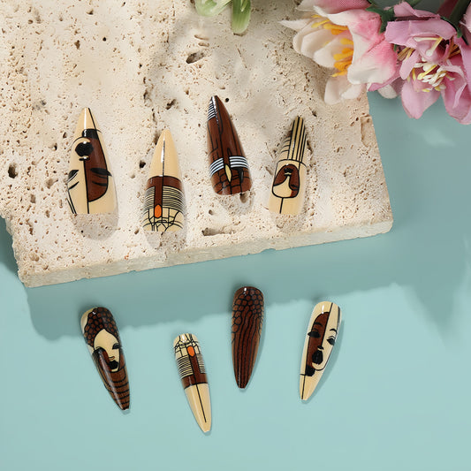 A set of long stiletto false nails with hand-painted tribal designs in beige, brown, and black colors displayed on a stone surface.