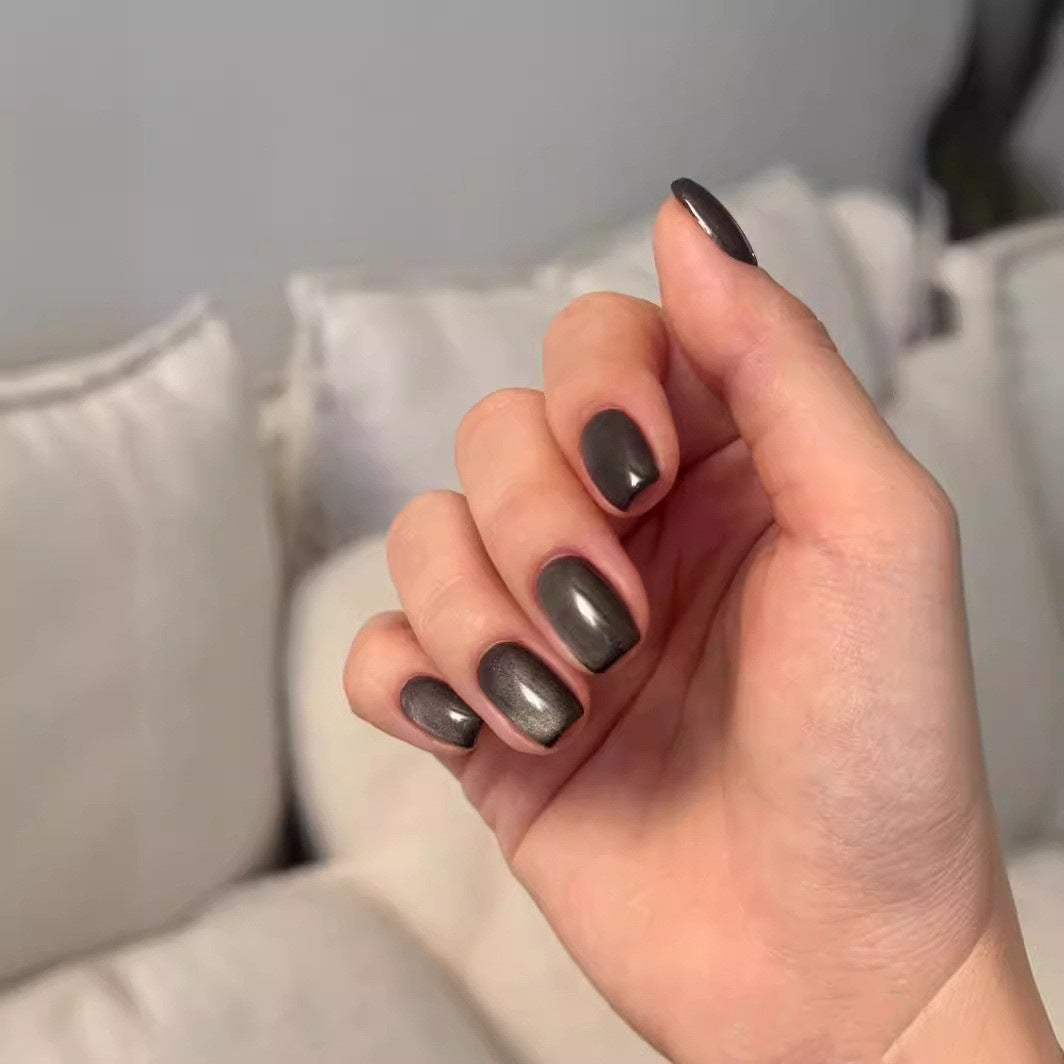 A hand showcasing press on nails in a short square shape with a charcoal grey color and metallic sheen finish.