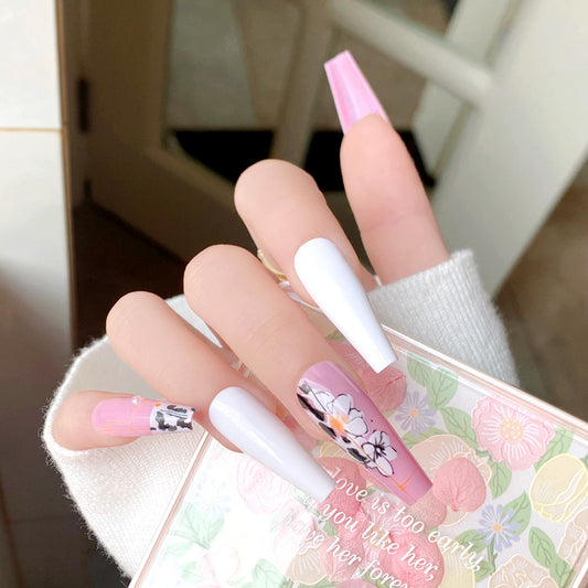 A set of long, press-on nails featuring a floral design with pink and white color scheme, displayed on a person's hands.