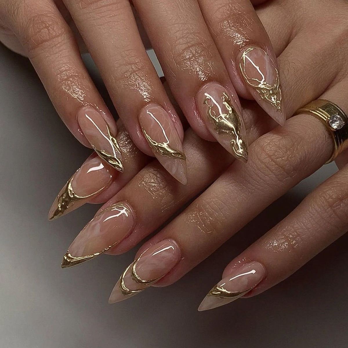 A set of long, stiletto-style beige press-on nails with metallic gold accents, displayed on a hand.