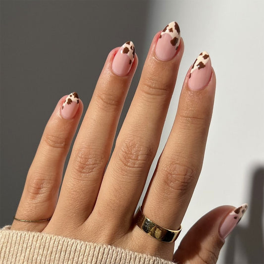 Hand with pink nails and cow print design wearing a gold ring on a neutral background