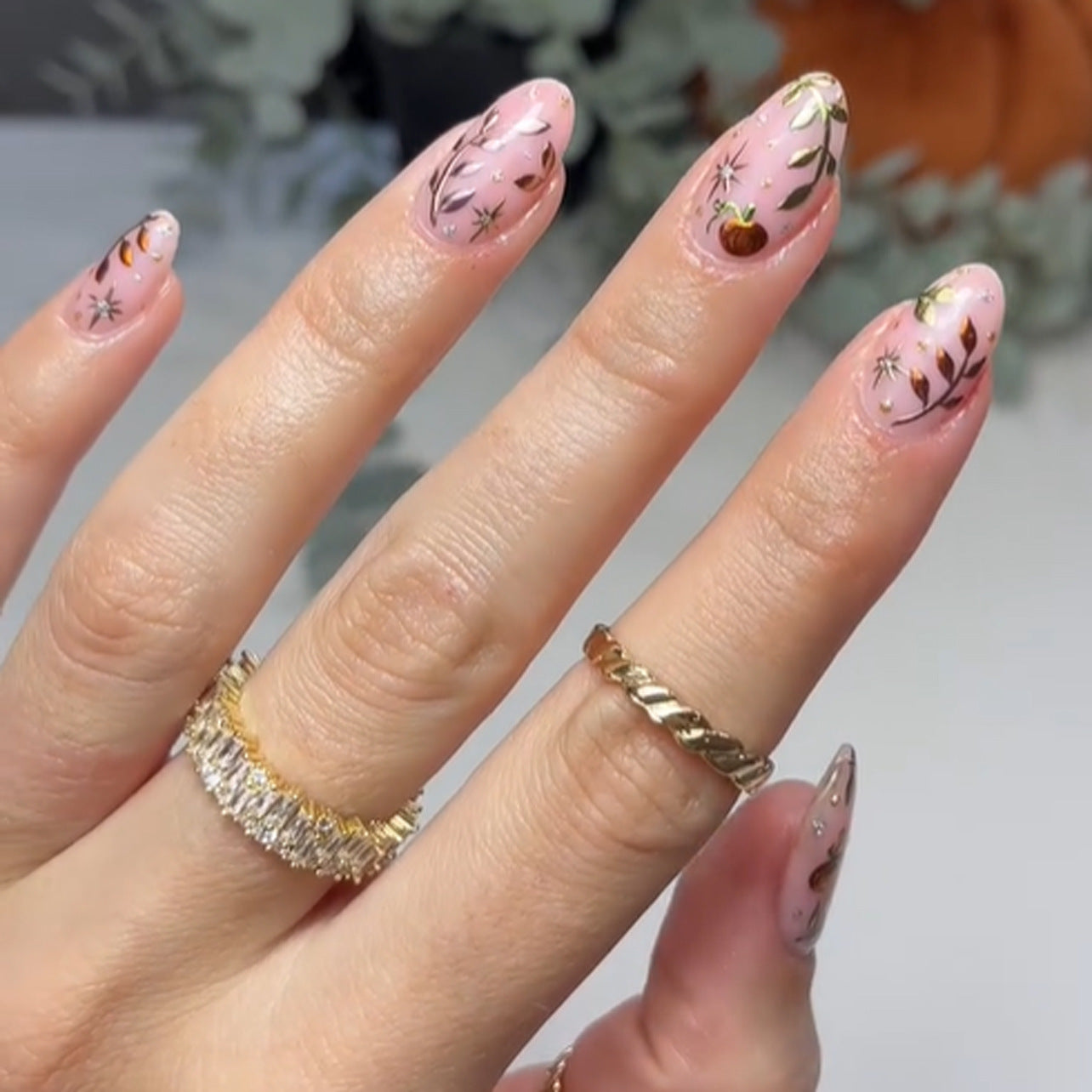 A close-up of a person's fingernails, which are painted with a medium almond pink color and feature gold floral accents.