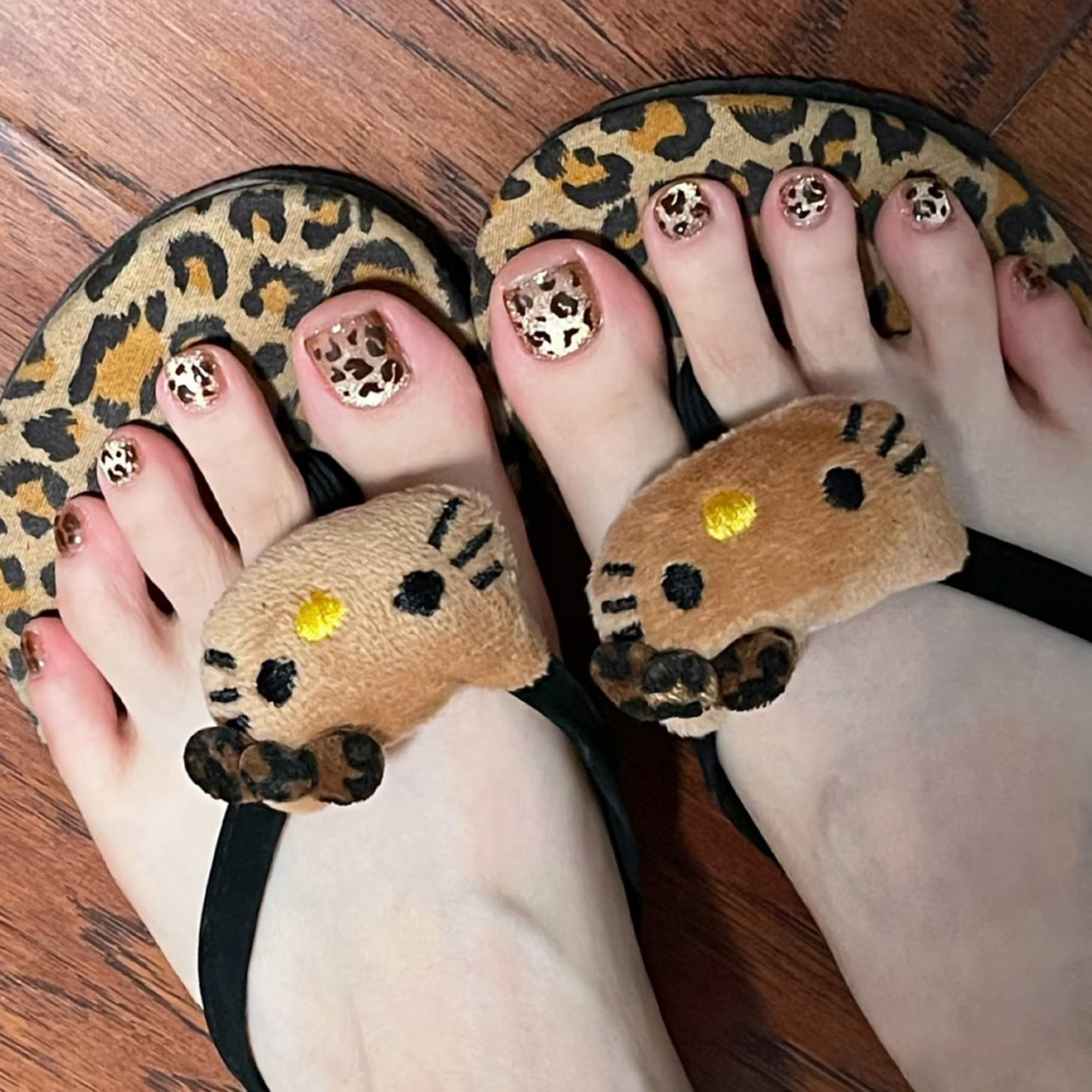 Feet wearing leopard print sandals with cat face design on a wooden floor.