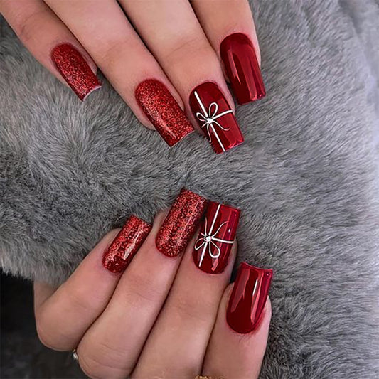 A set of long, square-shaped red press-on nails with glitter and a bow accent, displayed on a person's hands against a grey furry background.