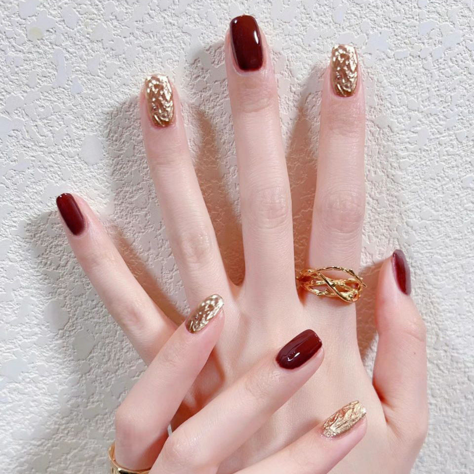 Hand with maroon nail polish and gold nail decorations wearing a gold ring on a textured white background