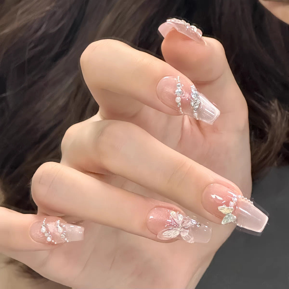 A set of long, coffin-shaped, pink press-on nails with floral charms and gemstone accents, displayed on a hand.