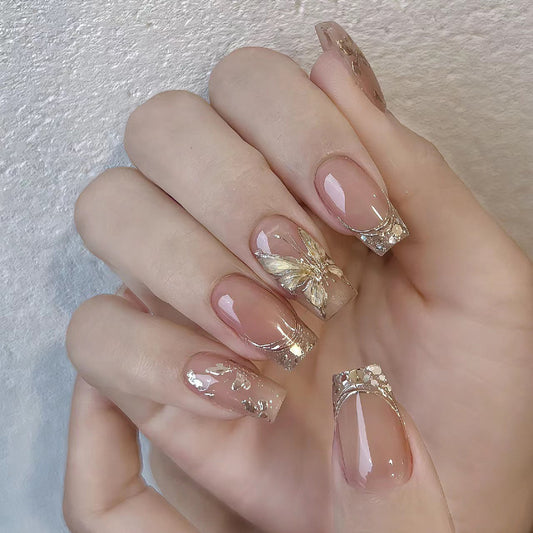 A hand with beige press-on nails featuring gold foil accents and a butterfly design at the tip of the nails.