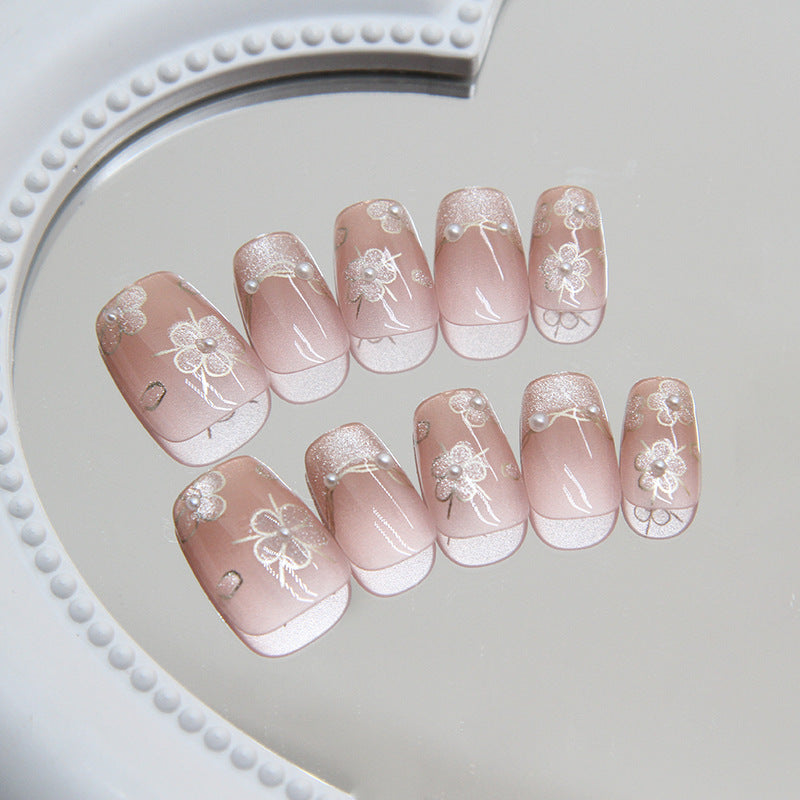 A set of pale pink press-on nails with glitter and floral accents, displayed on a reflective surface.