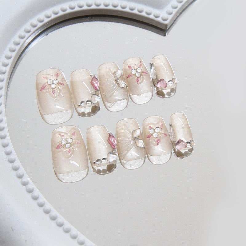 A set of beige false nails with floral patterns and rhinestone details, displayed on a white heart-shaped mirror.